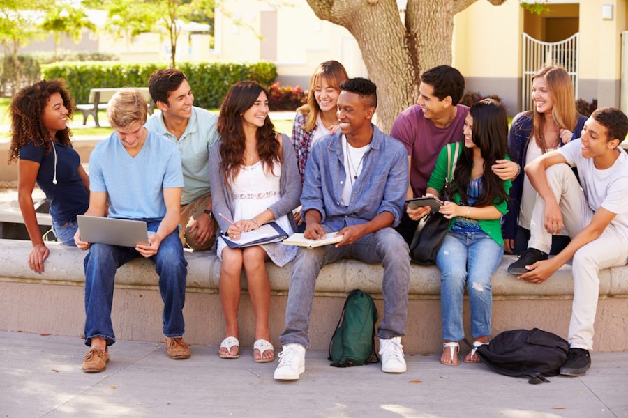 Outdoor Portrait Of High School Students On Campus Sitting Outside Holding Laptop, Digital Tablet and Text Book. Talking And Smiling To Each Other